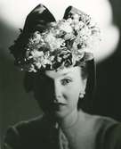Porträtt av kvinna i hatt dekorerad med blommor och rosett.