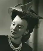 Porträtt av kvinna i hatt med flor och rosett, samt örhängen och halsband.