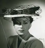 Porträtt av kvinna i halmfärgad stråhatt med marinblå fjäderbandsgarnering. Modell av Rose Valois.