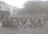 Gruppbild, många män från Landstormen iförda uniform(?) med armbindel , hattar och mössor fotograferade på en gräsmatta framför en stor byggnad. ( Kan vara Fästningen.) Männen har gevär med bajonett. Beställare: I. Lundin