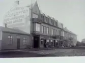 Affärsbyggnader i Falkenberg med reklam på husgavel med följande text: E. Malmgrens Möbelaffär; Bigge Johanssons Klädesaffär Manskläder Resefäkter m. m. Åt gatan finns skyltar för följande företag: Bigge Johansson Manufaktur- och Herrekiperings-affär; Fotografi-Atelier Sia Dahlberg; Gust. Westergren Speceriaffär; Damfrisering Stina Johansson.