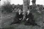 Två män sitter framför ett träd. Nils Gustavsson och Karl Fransen. I bakgrunden finns en stengärdsgård.