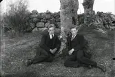 Två män sitter framför ett träd. Nils Gustavsson och Karl Fransen. I bakgrunden finns en stengärdsgård.