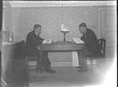 Interiör. Två män sitter vid ett bord och läser. Fotogenlampa.