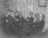 Kalas med folk som sitter utmed en vägg i salongen. En man röker och en yngling sitter i knät på en äldre kvinna, kanske hans mor.