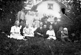 Kvinnor, män och barn sitter i gräset framför ett hus. Gustav Anderssons mor, Ottilia, nr 4 i övre raden och hans far, Johan August, nr 1 i mellanraden.