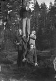Två stående män bärande en tredje stående man på sina axlar samt Gustav Andersson sittande framför dem i en barrskog.