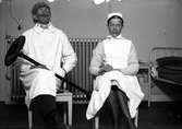 Två sittande män utklädda som läkare respektive sjuksköterska, i bakgrunden  ett element samt en sjukhussäng på Lungkliniken i Eksjö.