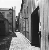 Rökerigränd i Jönköping, en kullerstensgata. Till höger ett bostadshus i trä med en port in till gården, ett antal fönster är öppna. I bakgrunden ligger ett stenhus utmed Odengatan.