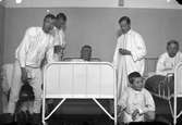 Fem män och en pojke, alla klädda i nattskjortor eller underkäder, vid två sjukhussängar. Fotografens anmärkning: 
