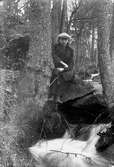 Gustav Andersson klädd i rock och keps, sitter på en sten vid en bäck i skogen.