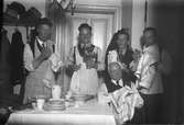 Fem män vid ett bord, fyra torkar glas med handdukar och en håller i en vas med blommor. På bordet finns bland annat en bricka, tallrikar och koppar. I bkagrunden syns en hatthylla med hattar och kläder.