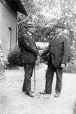 Två män står på en grusgång vid ett hus och skakar hand med varann, båda klädda i kostym och hatt och den ene med en promenadkäpp i handen.