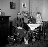 En äldre kvinnas högtidsdag. Hon sitter omgiven av blommor och en spettekaka står på bordet, telegram håller hon i handen.