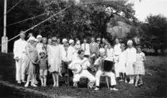 Sommarklädda kvinnor, män och barn. De två männen som sitter längst fram spelander banjo och dragspel. Hallen, Kållered, 1929-1950-tal.