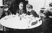 Bosgårdens barnträdgård 1938-1945. Barn som sitter vid ett bord utklädda till samer. Barnen har tillverkat sina egna enkla samekläder och mössor, med bl.a pärlträdning i lappfärger osv. Deras förskollärare Ingrid Hedlund hade gjort sitt examensarbete om samehantverk och denna kunskap använde hon i sitt pedagogiska arbete under flera år.