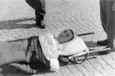Man ser en sjukling från ett koncentrationsläger 1945. Vederbörande ligger på en bår, som skall föras in i karantänsförläggningen i Kvarnbyskolan. Båren har ställts ned på Alléns stenläggning.