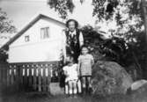 Modern Anne-Marie Lindholm, med barnen Marie-Louise och Benny, står i trädgården. I bakgrunden ses gaveln på Roten K 22, 1950-tal.