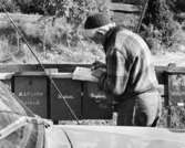 Konstnär Schmidtz hämtar sin post vid brevlådesamling i Skeppsdal. Bilåkande lantbrevbäraren Erik Johansson på linjen Åkersberga-Singö-Björnhuvud-Åkersberga.

Foto augusti 1961.