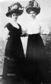 Två unga damer iklädda blommiga hattar och festkläder vid 1900-talet början.