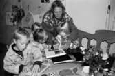 Fyra barn pysslar eller tittar i fotoalbum tillsammans med en vuxen person. Utställningsvernissage av och om Katrinebergs daghem på Mölndals museum 1993-09-10.