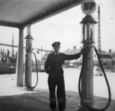 På Göteborgsvägen i Lackarebäck, snett emot lasarettet, låg denna BP bensinstation. Ville Nilsson står lutad mot en bensinpump. Kompisen Bror Eriksson jobbade också där. 1930-tal.