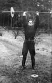 Givarens far Helmer Garthman, iklädd ridbyxor, står utomhus lyftandes en skivstång. Krokslätt, 1940-tal.