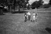 Fyra barn från Katrinebergs daghem står utomhus på en stor  inhägnad gräsmatta. Ett litet barn, iklädd hatt, väst och käpp, går mot fotografen. Ett par av barnen håller en stående träplanka. I bakgrunden ser man några hus samt höga träd.