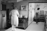 Christina Brussard och Kate Palm i köket på Katrinebergs daghem, 1992-93. Den ena lagar mat vid spisen och den andra står i en förlängd, vitt kaklad utbyggnadsdel och kontrollerar något. Båda kvinnorna bär förkläde.