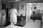 Christina Brussard och Kate Palm i köket på Katrinebergs daghem, 1992-93. Den ena lagar mat vid spisen och den andra står i en förlängd, vitt kaklad utbyggnadsdel och kontrollerar något. Båda kvinnorna bär förkläde.