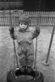 Lille Fred, ca två år, står på en gunga gjord av däck. Han har overall, mössa och vantar på sig. Marken är sandig och omgärdad av staket. Lunkentussen, Katrinebergs daghem 1992.