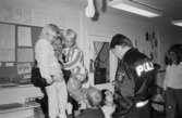 Katrinebergs daghem har besök av två manliga poliser. Dessa är omringade av flera barn varav den ene polisen håller två barn i famnen. Deras fröken står lite vid sidan av. Alla ser mycket glada ut och barnen tittar med stor beundran på männen.