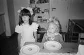 Två flickor står inomhus bakom ett bord på Katrinebergs daghem. På bordet står två tallrikar med var sin kaka på. Båda flickorna har dekoration i håret. I bakgrunden står en hylla innehållande pysselprylar.