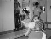 Fyra små barn sitter/står bredvid daghemmets städerska. Ett barn pratar med henne. Vid sidan om står städvagnen. I bakgrunden syns en personal som ordnar i köket. Katrinebergs daghem, 1992.