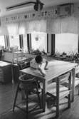 I köket på Katrinebergs daghem. En pojke har rest sig från sin trip trap stol och sträcker sig över köksbordet av furu. Han torkar med en trasa på bortre delen av bordet. I bakgrunden ser man fönster med gardiner. Ovanför hänger tavlor av versaler i bokstavsordning.