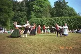 Ett folkdanslag som dansar omkring på Gunnebo slotts gräsmattor. Några sitter runt om och beskådar dansen.