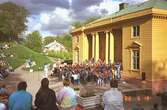 Föreställning med flertalet barn som spelar och sjunger. De står på trappan till Gunnebo slotts framsida. På grusplanen framför står eller sitter människor och ser på.