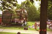 Människor sitter på gräset och lyssnar till Lasse Berghagen, som sjunger på en tillfälligt uppbygd scen på Gunnebo slotts baksida, 29 maj 1988.