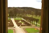 En del av parken på Gunnebo slotts högra baksida. Bilden är tagen inifrån och ut genom ett öppet brunt fönster. I bakgrunden skymtar man dammen.