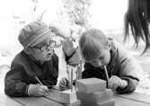 Två barn sitter och pysslar vid ett bord utomhus. Holtermanska daghemmet juni 1973.