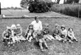 Några barn och en fröken sitter i gräset. Holtermanska daghemmet 1973.