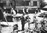 En man står på en gårdsplan bredvid några cyklar. I bakgrunden ses barn och vuxna som packar/packar ur en folkvagnsbuss framför ett stort hus. 1950-1960-tal.