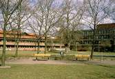I förgrunden ser man ett par gula bänkar, en papperskorg och några träd som står i Stadshusparken. I bakgrunden ses Mölndals stadshus, mars 1993.