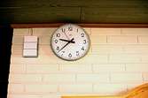 En klocka uppsatt på en tegelvägg, troligen från Sagåsen 1980-talet. Till vänster om klockan sitter en ringklocka.