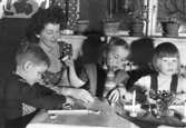 Bitr. föreståndare Margit Emilsson (gift Wannerberg -52) sitter tillsammans med två syskon och ett annat barn i 
New look, egenhändigt ritade kläder, Krokslätts daghem år 1949.

Margits rödprickiga förkläde är av samma tyg som gardinerna bakom henne. Tyget är från Mölnlycke fabriker.
