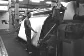 Denny Andersson står vid en maskin för kartongtillverkning i KM 3, Byggnad 6. 
Bilden ingår i serie från produktion och interiör på pappersindustrin Papyrus, 1980-tal.