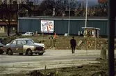 Domus barack vid Nya Torget i Mölndal, 1970-tal. Främst ses parkerade bilar uppställda på en parkeringsplats. Till höger ses en liten kiosk.