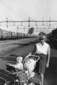 Berit Gustafsson med dottern Ingela i barnvagn cirka 1968/69. De står vid en tågstation, troligtvis i Lindome.