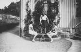 Sommar i Lindome i början av 1940-talet. En ung kille balanserar ståendes på en bräda. På var sida av brädan sitter två unga tjejer. I bakgrunden ses en villa och en trädgård.