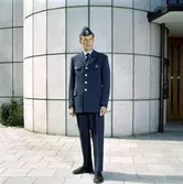 Brevbärare fotograferad utomhus i 1962 års vinteruniform. Uniformen är av gråblått ylletyg; enradig kavaj med fyra stora uniformsknappar, fyra fickor med lock. Bröstfickans lock knäpps med uniformsknapp av mellanstor storlek. Byxor utan slag och båtmössa försedd med mössmärke med en vinkel vilket var avsett för postiljoner och expeditionsvakter.